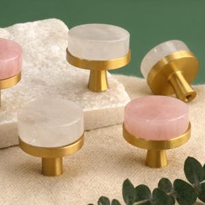 Puede incluir: Un conjunto de tiradores decorativos para armarios. Los tiradores presentan una parte superior redonda de piedra preciosa pulida, en rosa o transparente, sobre una base dorada. Los tiradores se muestran sobre una piedra blanca y un fondo de tela beige.