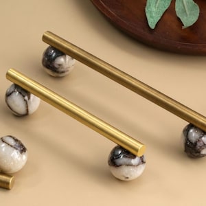 May include: A set of cabinet pulls featuring long, cylindrical brushed brass bars attached to spherical bases made of white stone with dark brown veining. The hardware components are arranged on a flat surface, showcasing the metallic finish.