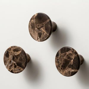 May include: Three circular cabinet knobs featuring a polished dark brown marble surface with intricate light brown veining. Each knob has a smooth, rounded face and a cylindrical base, showcasing natural stone patterns in a set of three.