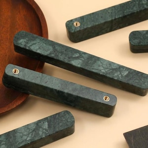 May include: A collection of rectangular cabinet pulls crafted from dark green marble with a natural veined pattern. Each handle features two circular brass-toned metal inserts for mounting, showcasing a smooth, polished finish and rounded edges.