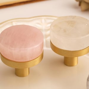 Peut inclure: Deux boutons de meuble décoratifs. L'un est en quartz rose clair, l'autre en quartz clair. Les deux ont une base et une tige dorées. Les boutons sont sur une assiette blanche à motifs floraux.