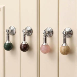 May include: Four drop-style cabinet pulls featuring polished chrome hardware and spherical stone accents. The pulls include a dark green marble, a dark brown marble, a pale pink rose quartz, and a light beige stone, all mounted on cream doors.