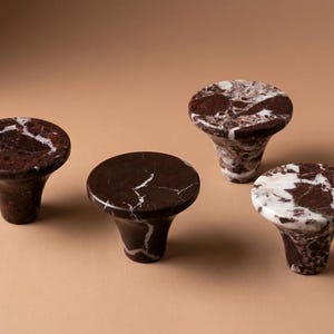May include: Four cabinet knobs crafted from dark brown and white marble. Each knob features a smooth, circular top surface that tapers down to a narrower base. The natural stone displays unique veining patterns in shades of deep chocolate brown and bright white, creating a polished, reflective finish across the set of hardware.