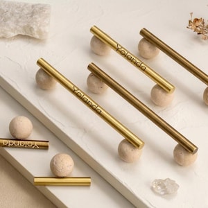 May include: A collection of gold-toned cabinet pulls with the name "Yolanda" engraved on them. Each pull is mounted on a small, round, beige stone. The pulls are arranged on a white, textured surface.
