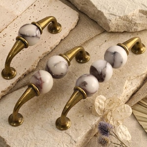 May include: Three cabinet handles featuring antique brass finish metal bases and spherical white marble accents with grey veining. The handles vary in length, with one featuring a single marble sphere, one with two, and one with three spheres.