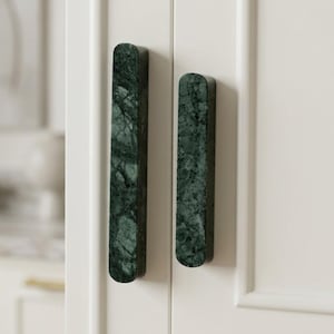 May include: Two vertical cabinet handles made of dark green marble with natural white veining. The handles have a rectangular shape with rounded ends and are mounted on a white cabinet door, featuring a smooth polished stone surface texture.