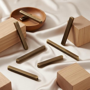 May include: Several rectangular, antique brass cabinet pulls are displayed on a cream satin surface. Wooden blocks and a wooden bowl are also present. The word "Yolanda" is visible on the bowl.