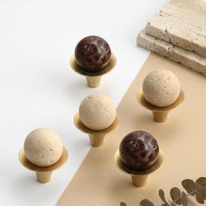 May include: A collection of decorative cabinet knobs. The knobs feature a brass base with a round, stone-like sphere on top. Some spheres are light beige, while others are a dark reddish-brown. The knobs are arranged on a neutral background.