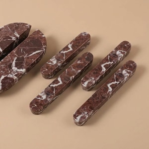 Può includere: Una collezione di oggetti in marmo rosso scuro con venature bianche, tra cui un pezzo semicircolare e quattro barre rettangolari allungate con estremità arrotondate, tutti caratterizzati da una texture in pietra lucida con motivi naturali.