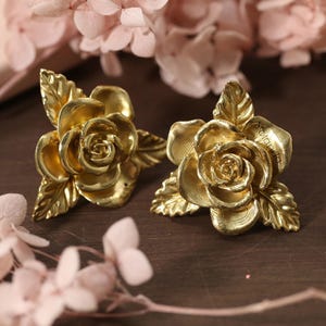 May include: Two gold-colored rose-shaped earrings with detailed petals and leaves. The earrings are on a dark brown surface, with pink flowers in the background. The earrings have a vintage aesthetic.