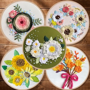 May include: Five embroidery hoops with different floral designs. The hoops are made of wood and have a white background. The designs include a pink rose, white daisies, yellow sunflowers, orange flowers, and a mix of pink, white, and blue flowers.