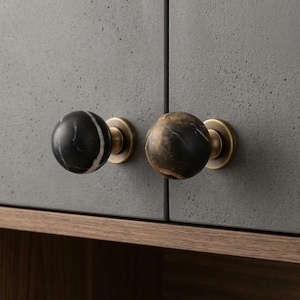 Può includere: Due pomelli sferici per armadi montati su ante grigie testurizzate. Il pomello sinistro è in marmo nero con venature bianche, quello destro è in marmo marrone e nero. Entrambi i pomelli presentano una base circolare color ottone e un gambo.