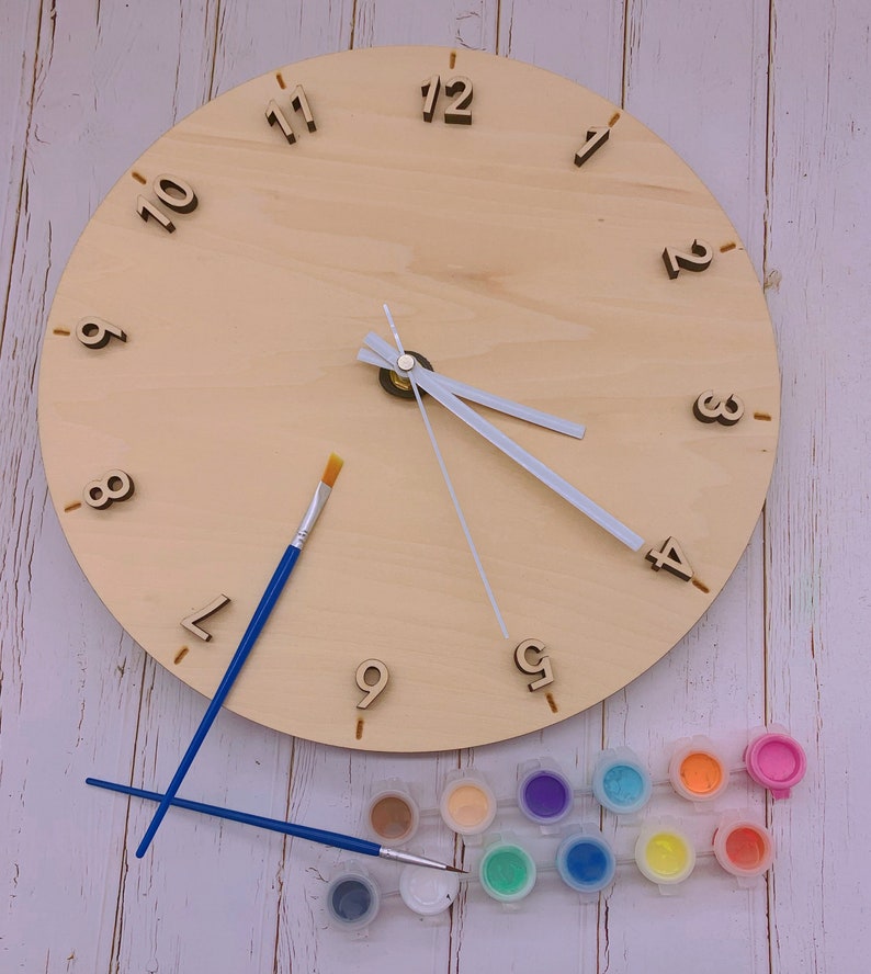 May include: A wooden clock face with Roman numeral markings and a set of paint pots and paintbrushes. The clock face is blank and ready to be painted.