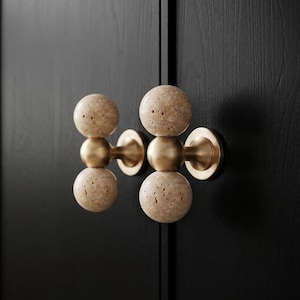 May include: Two cabinet knobs featuring a vertical arrangement of two porous, beige travertine stone spheres connected by a central brushed brass metal sphere, mounted on a circular brass base attached to a dark black wooden cabinet door surface.
