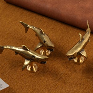 Puede incluir: Tres figuras de tiburones de latón con aletas detalladas y bocas abiertas, cada una montada sobre una pequeña base redonda. Los tiburones están dispuestos sobre una superficie marrón, mostrando su brillo metálico.