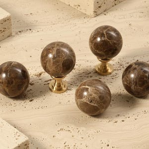 Op de afbeelding: Vijf decoratieve kastknoppen. De knoppen zijn bolvormig, gemaakt van bruin marmer met unieke adering, en gemonteerd op goudkleurige voetjes. De knoppen worden gepresenteerd op een lichtbeige oppervlak met rechthoekige stenen accenten.