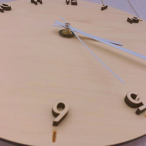May include: A wooden clock face with black numbers and white hands. The clock face is light brown and has a wood grain texture.