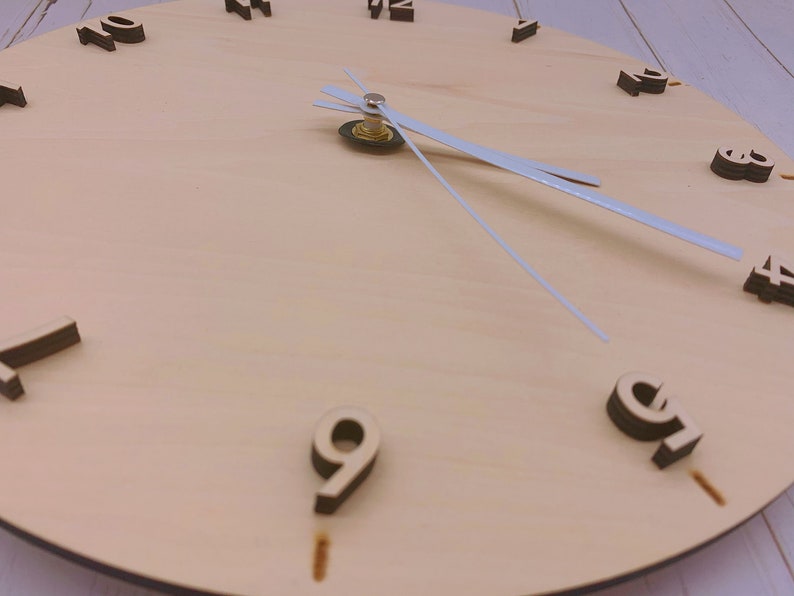 May include: A wooden clock face with black numbers and white hands. The clock face is light brown and has a wood grain texture.