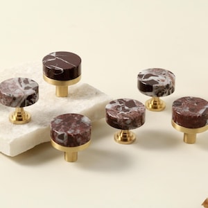 Może przedstawiać: Zestaw sześciu gałek do szafek. Każda gałka ma okrągłą, ciemnoczerwoną i białą marmurową górę, osadzoną na złotej podstawie. Gałki są ułożone na jasnej powierzchni, odpowiednie do dekoracji wnętrz.