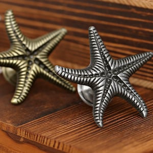 Könnte beinhalten: Zwei sternförmige Schrankknöpfe, einer bronzefarben und einer silberfarben, mit detaillierten Rippen. Sie befinden sich auf einer Holzoberfläche, was auf ein maritimes Thema hindeutet. Ideal für Schubladen.