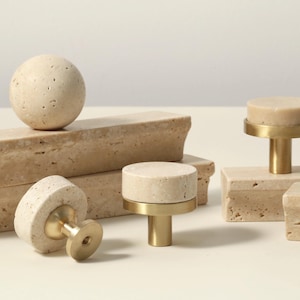 Könnte beinhalten: Eine Sammlung beigefarbener Stein-Schrankknöpfe und -Griffe mit Messingbeschlägen. Die Knöpfe sind rund, zylindrisch und kugelförmig. Die Griffe sind rechteckig. Die Beschläge sind goldfarben.