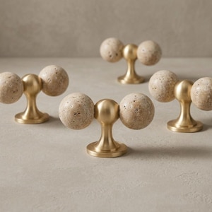 Può includere: Un set di quattro pomelli per armadietti, ciascuno caratterizzato da un perno centrale in metallo dorato con base circolare svasata. Ai lati di ogni perno sono fissate due sfere in pietra beige porosa, creando un design simmetrico.