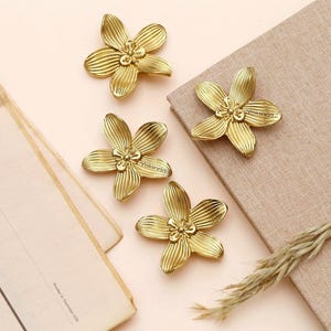 Könnte beinhalten: Vier goldfarbene, blütenförmige Dekorationsgegenstände sind auf einer beigen Oberfläche angeordnet. Die Blüten haben detaillierte Blütenblattmuster und einen zentralen floralen Akzent. Eine Zeitung und ein Zweig trockenes Gras sind ebenfalls vorhanden.