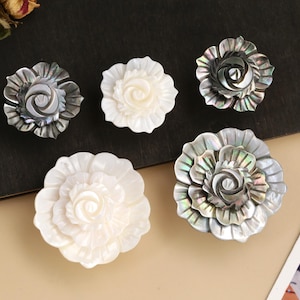 May include: An assortment of flower-shaped decorative elements. The elements are in shades of white and iridescent gray, with detailed petal designs. The items are arranged on a dark surface with a light-colored background.