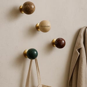 Peut inclure: Ensemble de patères murales décoratives avec des sphères en marbre de différentes couleurs, montées sur des bases en laiton. L'une des patères porte le nom "Yolanda". Un sac fourre-tout beige est suspendu à un crochet vert.