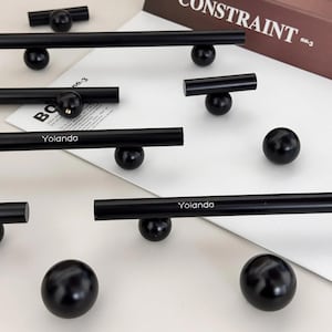 May include: Black cabinet hardware featuring cylindrical handles and spherical knobs. Some handles have the name "Yolanda" printed on them. The hardware is displayed on a white surface with a book in the background.
