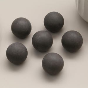 May include: Six dark gray, spherical objects are arranged on a light gray surface. The spheres have a matte finish and appear to be made of a solid material. A decorative white plate and a white bowl are partially visible in the background.