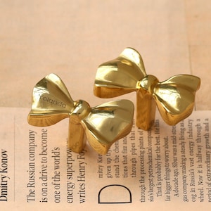 May include: Two gold-coloured bow-shaped cabinet knobs. The knobs are made of a shiny metal and are positioned on a newspaper background. The word "Yolanda" is engraved on one of the bows.