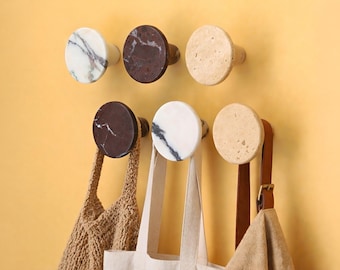 Può includere: Sei ganci da parete circolari disposti su due file. La fila superiore presenta marmo bianco con venature grigie, marmo marrone scuro con venature bianche e pietra beige. La fila inferiore sostiene una borsa in maglia beige, una borsa in tela bianca e una borsa in pelle marrone.