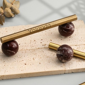 May include: A set of brass-toned metal rods and dark maroon marble spheres. One rod is engraved with the name Yolanda. These components are arranged on a rectangular, porous, beige stone tile with a textured surface and visible natural indentations.