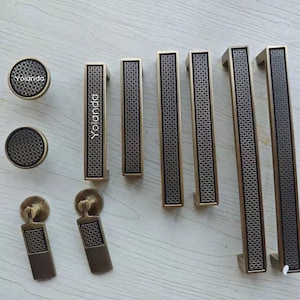 Può includere: Una collezione di ferramenta per mobili color bronzo, tra cui pomelli, maniglie e tiranti. La ferramenta presenta un motivo a rete geometrica e il marchio "Yolanda". Il set include varie dimensioni e stili.