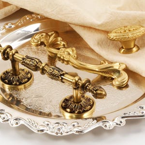 Può includere: Una collezione di ferramenta in ottone ornate, tra cui maniglie per cassetti e un pomello per armadietto, esposte su un vassoio d'argento. La ferramenta presenta intricati disegni floreali e a volute, con un'estetica vintage.