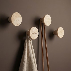 Op de afbeelding: Vier beige stenen wandhaken van verschillende formaten zijn op een donkerbruine muur gemonteerd. Eén haak houdt een lichtgekleurde handdoek vast en een andere heeft een bruine leren riem eraan hangen. Het woord "Yolanda" staat op een van de haken.