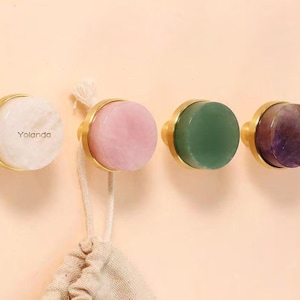 Może przedstawiać: Cztery dekoracyjne gałki do szafek ze złotymi podstawami i kolorowymi kamiennymi blatami. Gałki są białe, różowe, zielone i fioletowe. Na pierwszej gałce wydrukowano słowo "Yolanda".