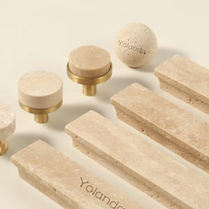 Könnte beinhalten: Eine Sammlung von Schrankbeschlägen, darunter Knöpfe und Griffe. Die Knöpfe haben eine hellbeige, kreisförmige Oberseite mit einem Messingfuß. Die Griffe sind rechteckig und haben eine ähnliche beige Farbe. Das Wort "Yolanda" ist auf einigen Teilen eingraviert.