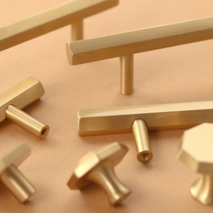 May include: A set of six gold-toned cabinet hardware pulls and knobs. The pulls are rectangular with a hexagonal shape and the knobs are round with a hexagonal shape.