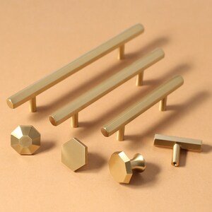 May include: A set of gold-toned cabinet hardware, including pulls and knobs, in various shapes and sizes. The hardware is arranged on a light brown surface.