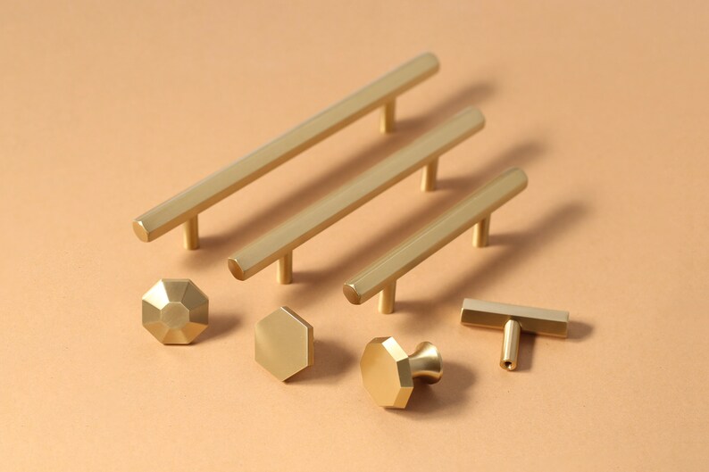 May include: A set of gold-toned cabinet hardware, including pulls and knobs, in various shapes and sizes. The hardware is arranged on a light brown surface.