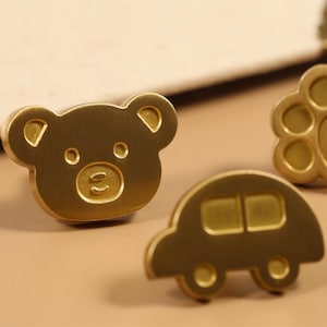 Peut inclure: Trois boutons de meuble dorés en forme de tête d'ours, de voiture et d'empreinte de patte. Le bouton en forme d'empreinte de patte porte le nom "Yolanda". Ces boutons sont pour les meubles.