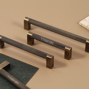 Może przedstawiać: Cztery uchwyty do szafek w odcieniu brązu z teksturowanym, ciemnoszarym środkiem i prostokątnymi końcami. Słowo "Yolanda" jest wydrukowane na dwóch uchwytach. Uchwyty są ułożone na neutralnym tle.