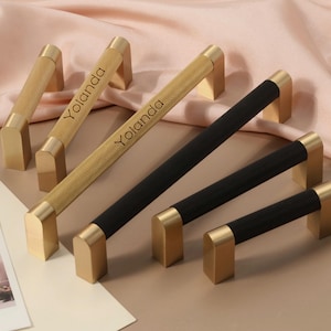 Könnte beinhalten: Eine Auswahl an goldfarbenen Schrankgriffen, einige mit strukturiertem Muster und schwarzen Akzenten. Der Name "Yolanda" ist auf einigen eingraviert. Die Hardware wird auf einem pfirsichfarbenen Stoff präsentiert, was auf ein Wohndekor-Thema hindeutet.