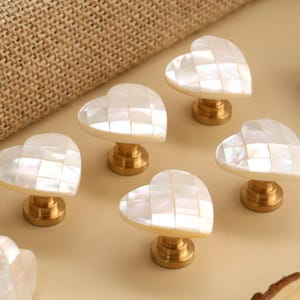 Può includere: Pomelli per armadi a forma di cuore con un design a mosaico in madreperla. I pomelli hanno una base color oro. I pomelli sono disposti su una superficie beige chiaro. Ferramenta decorativa per mobili.