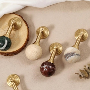 Op de afbeelding: Een verzameling decoratieve kastknoppen met messing hardware en stenen bollen in verschillende kleuren, waaronder groen, beige en bordeaux. Geschikt voor interieurdecoratie.