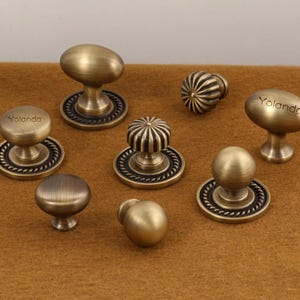 May include: A selection of antique brass cabinet knobs in diverse shapes, such as oval, round, and ribbed. Each knob is mounted on a decorative base with a rope-like border. The name "Yolanda" is engraved on some of the knobs.