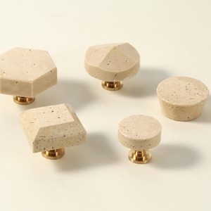 Puede incluir: Cinco tiradores decorativos para armarios en varias formas geométricas: un hexágono, una forma facetada, un cuadrado, un cilindro y un disco pequeño. Cada tirador es de color beige claro con una textura moteada y una base de latón.