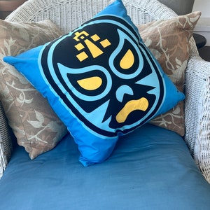 May include: A blue cushion with a black and yellow wrestling mask design. The cushion is sitting on a white wicker chair with a brown patterned cushion.